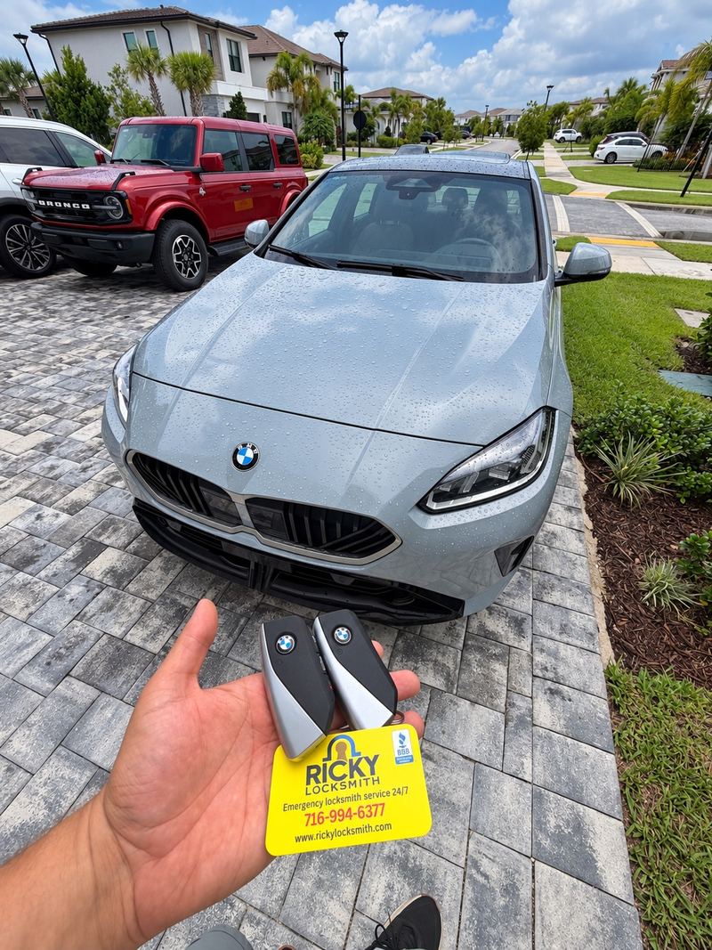 Two new BMW keys and a Ricky Locksmith card held in front of a gray BMW